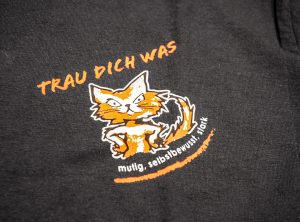 Tshirtdruck: Trau-Dich-Was Logo auf verschiedenfarbigen Tshirts.