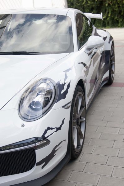 Weißer Porsche GT3 von schräg vorne, halb im Bild zu sehen mit einer schwarz-grauen Streifenbeklebung in Fetzenoptik.