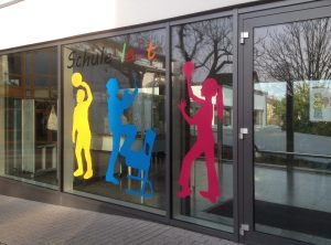 Farbenfrohe Kinderumrisse aus Folie und der Slogan "Schule lebt" an den Fenstern der Oberasbacher Grundschule.