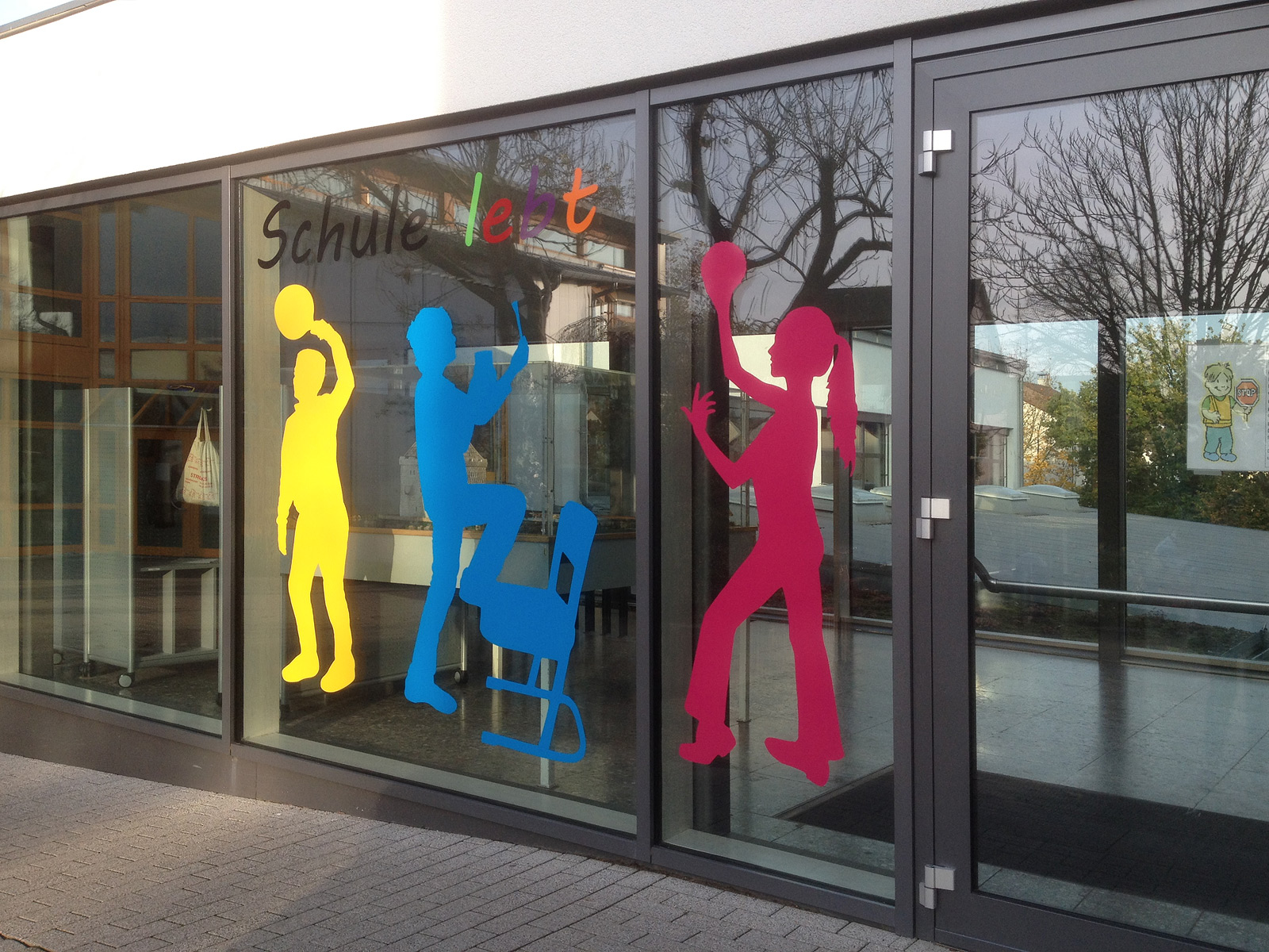 Farbenfrohe Kinderumrisse aus Folie und der Slogan "Schule lebt" an den Fenstern der Oberasbacher Grundschule.